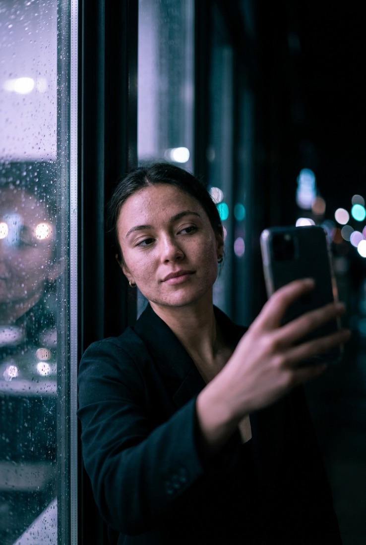 A young woman taking a selfie by a window at night