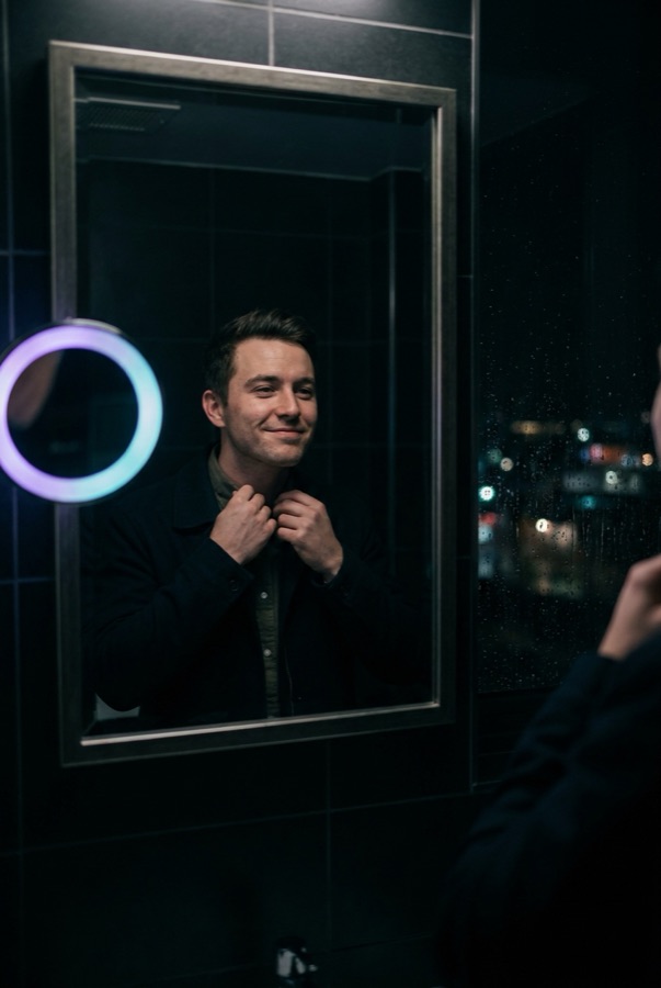 A man checking his collar in a mirror lit by a ring light at night